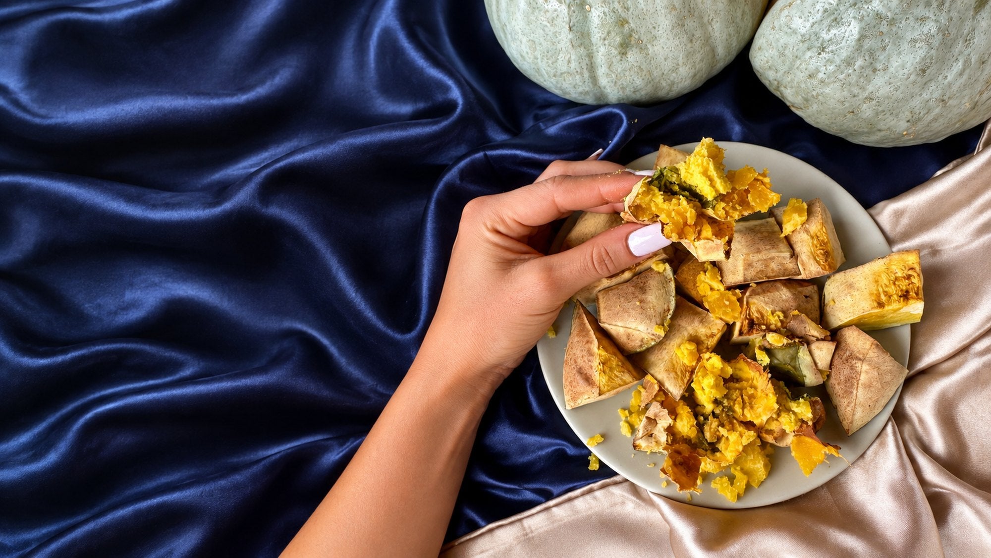 Una mano prende un pezzo di zucca al forno da un piatto, accanto due zucche intere